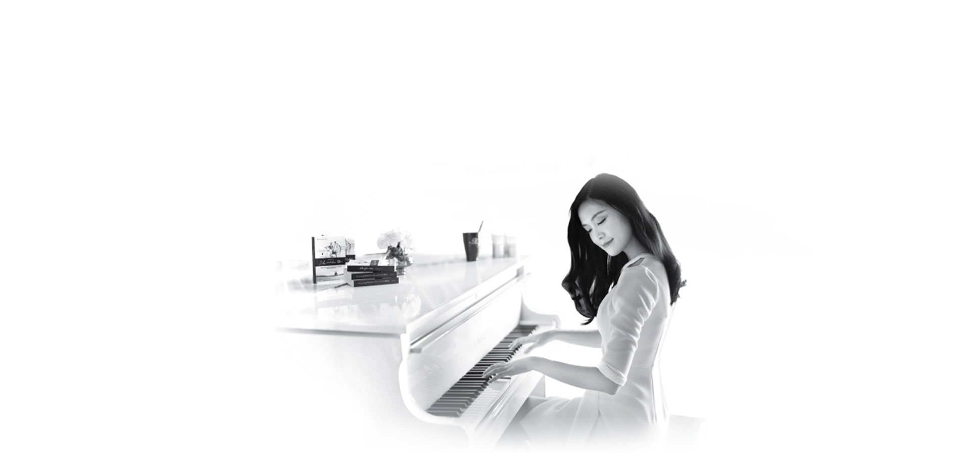 A woman in white plays grand piano beside a lake — coffee, books, and flowers on the piano surface